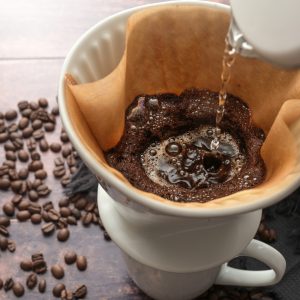 Brewed coffee, pouring hot water on ground coffee in a filter on a mug for a stimulating drink, rustic wooden table with some beans, copy space, selected focus, narrow depth of field